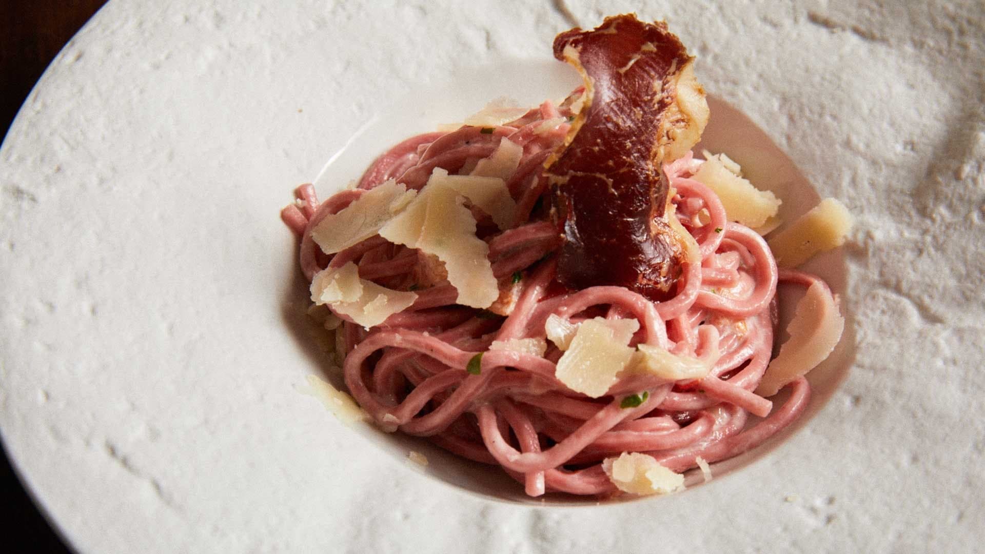 Pancar rengi spagetti, ince parmesan dilimleri ve kıtır bir dilim füme etle beyaz tabakta sunulmuş.