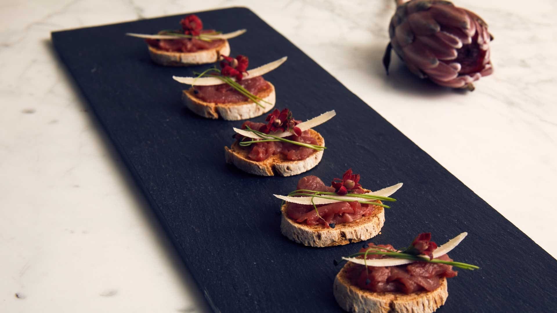 Kavrulmuş ekmek dilimleri üzerine et tartarı, parmesan tıraşlaması ve kırmızı çiçeklerle siyah taş tabakta sıra halinde dizilmiş kanepeler.