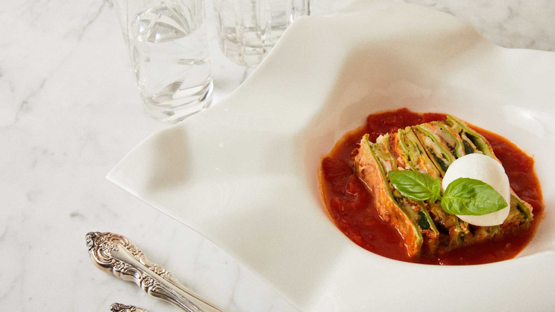 Kabak dilimli bir lazanya dilimi domates sosu içinde, üzerinde mozzarella topu ve fesleğen yaprağıyla tabaklanmış.