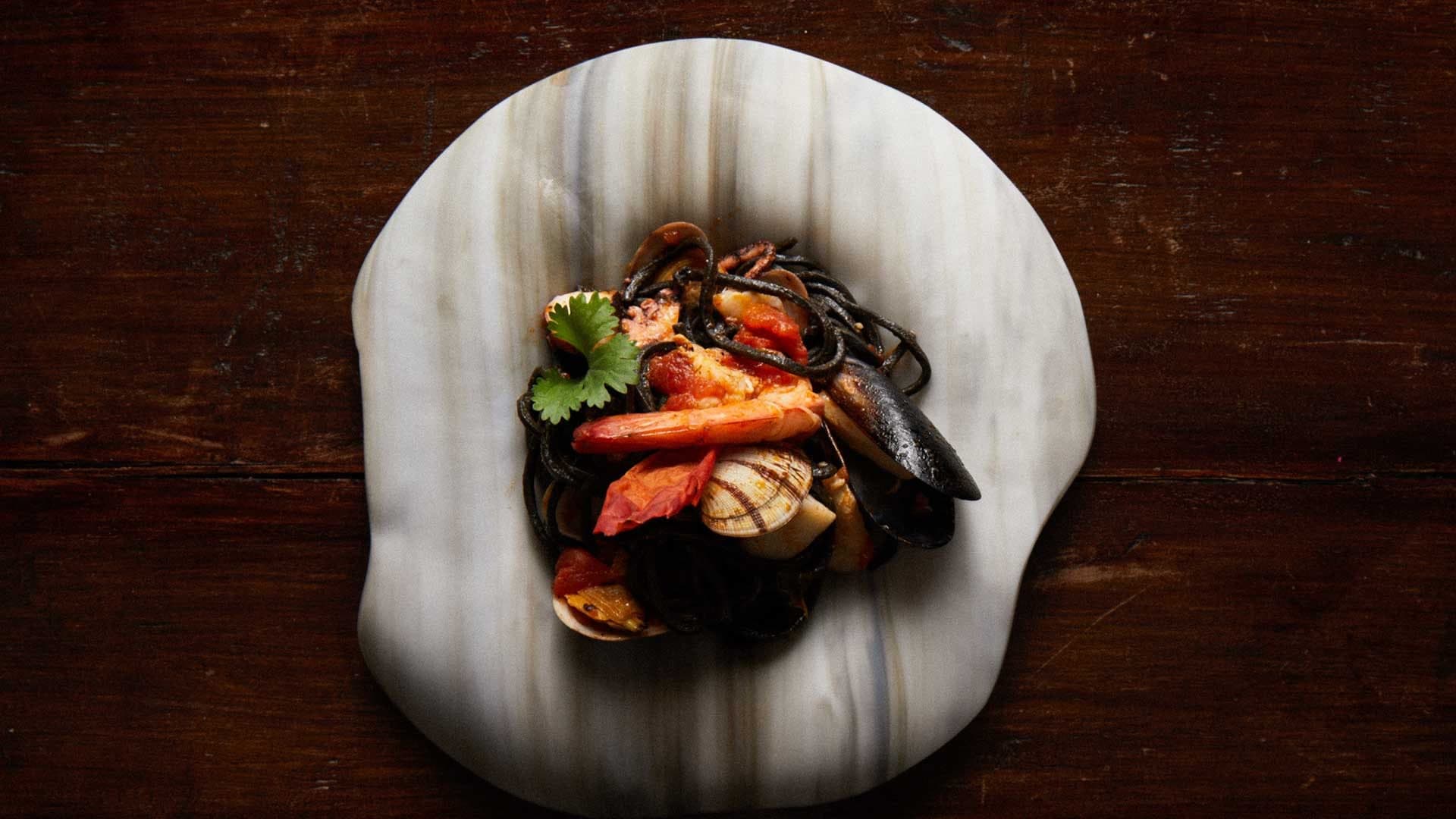 Siyah mürekkepli spagetti; karides, midye, kum midyesi ve kişnişle mermer tabakta, koyu ahşap masada üstten çekilmiş.